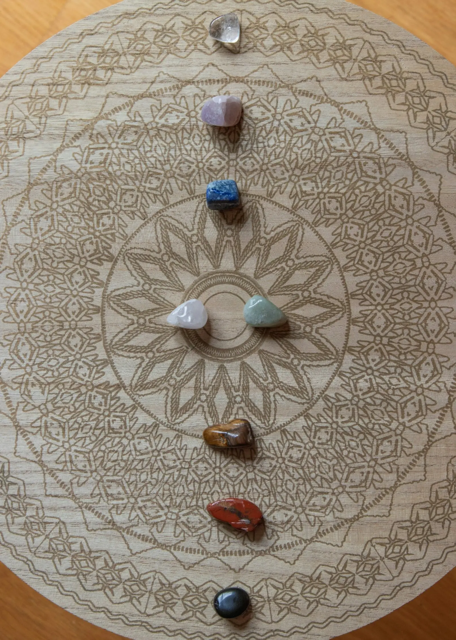 Eight polished stones in different colours lie in a vertical row on a wooden disc engraved with a mandala.